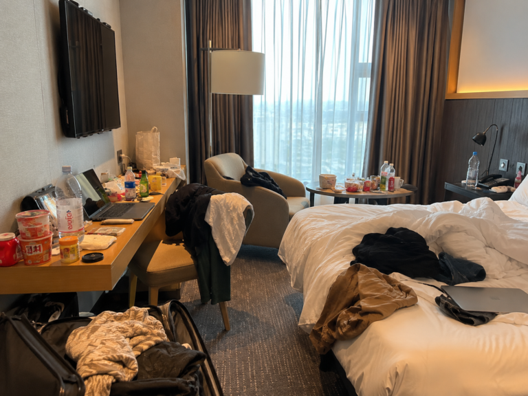 Messy hotel room showing long-term hotel living with clothes, food containers, luggage, and a laptop on the desk.
