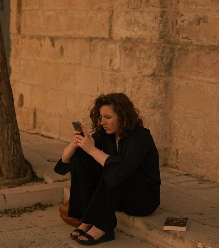 Young woman sitting alone outdoors while looking at her phone, representing modern loneliness, texting culture, and digital communication