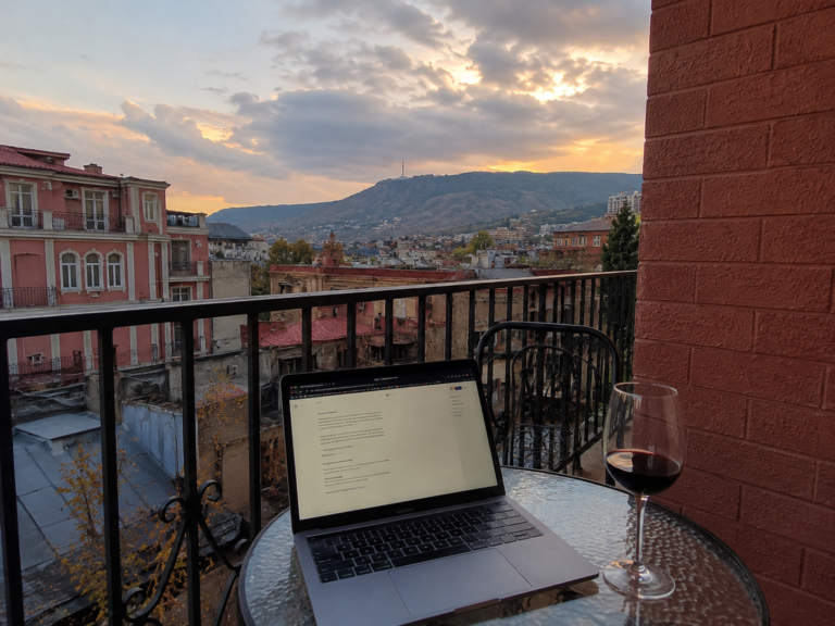 Digital nomad working remotely from a balcony in Tbilisi, Georgia, with a laptop and glass of wine overlooking the city at sunset