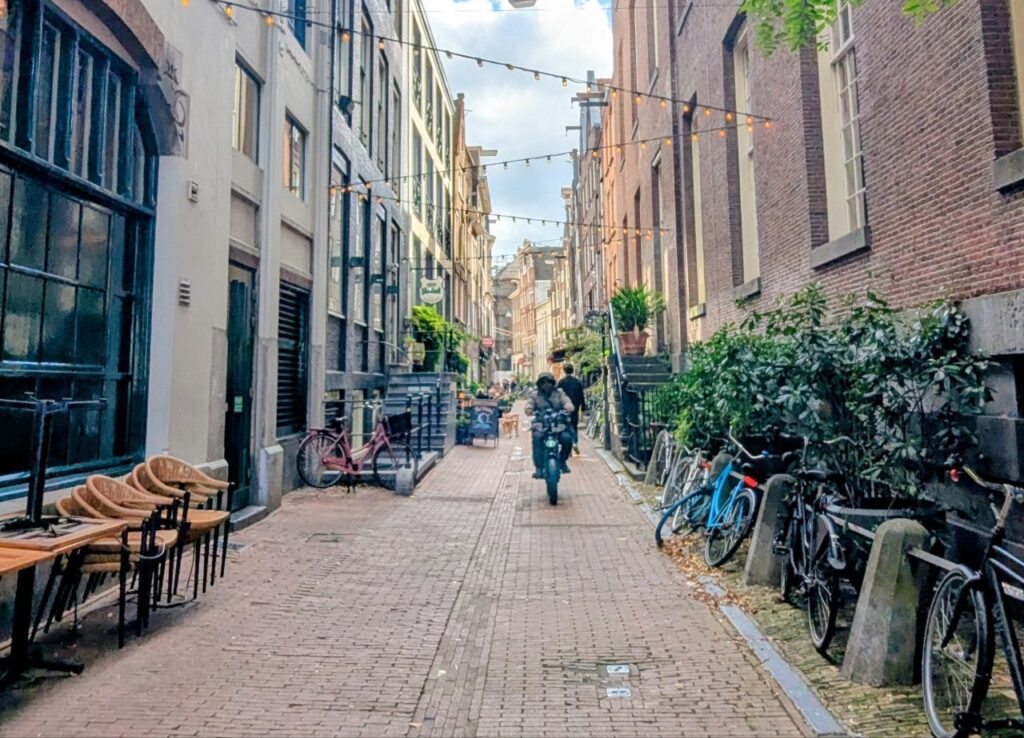 Narrow Amsterdam residential street with bicycles, outdoor seating, historic buildings, and local residents cycling through the neighborhood.