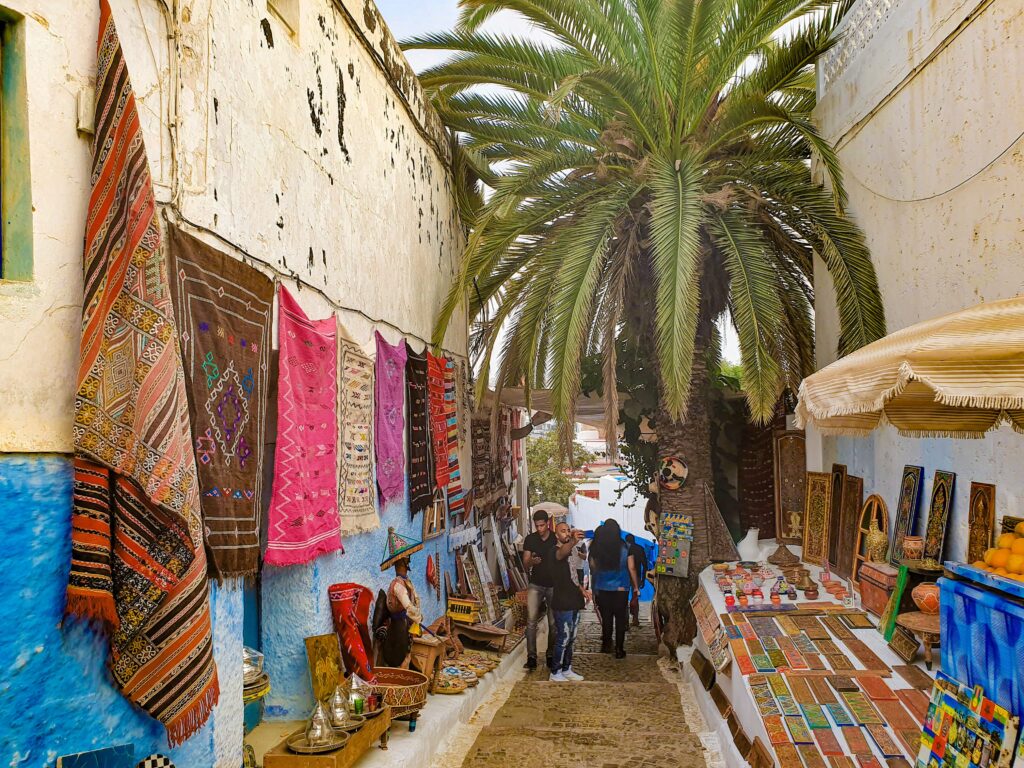 Traditional market street in Chefchaouen, Morocco, with local handicrafts, colorful rugs, and palm trees reflecting Moroccan culture.