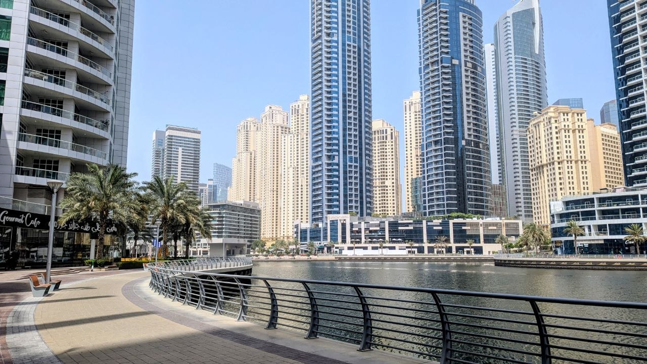 Dubai Marina waterfront on a sunny summer day with modern skyscrapers, palm trees and a nearly empty pedestrian promenade during the hottest season.