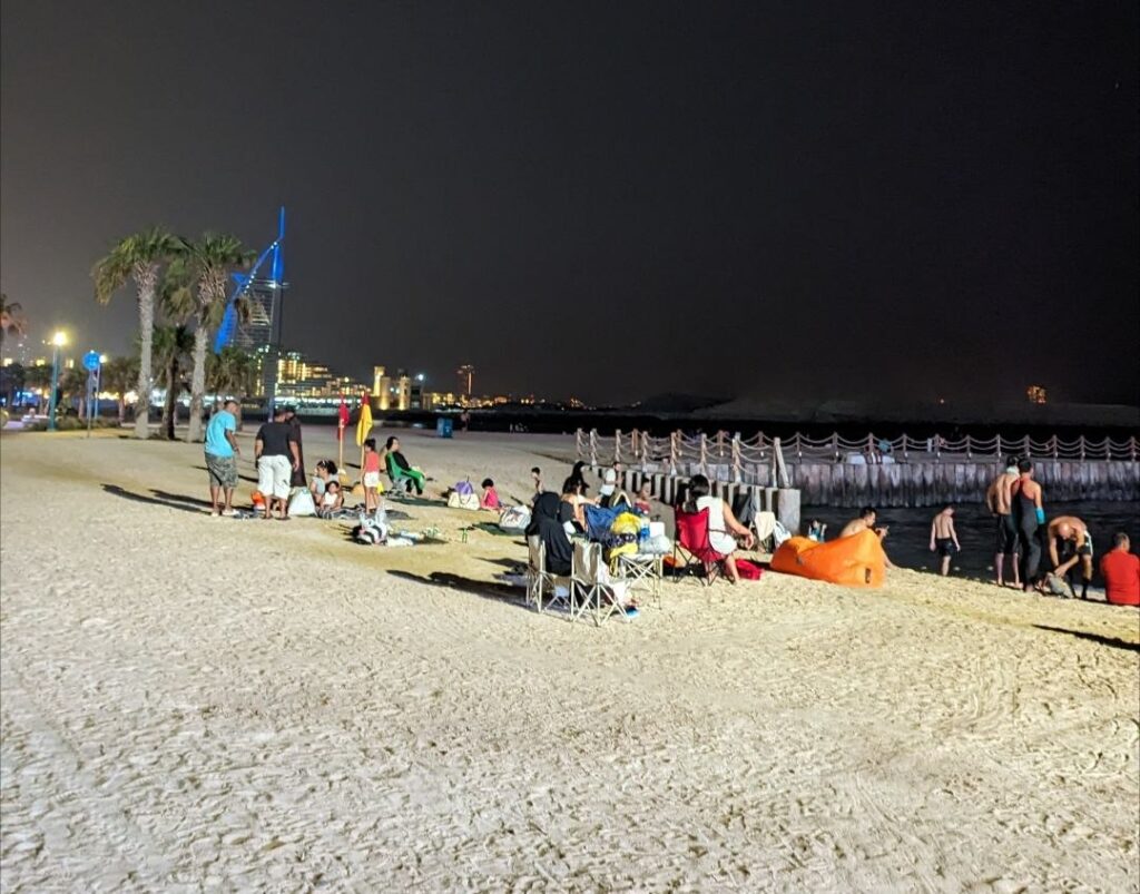 People relaxing on Kite Beach in Dubai at night during summer with the Burj Al Arab illuminated in the background